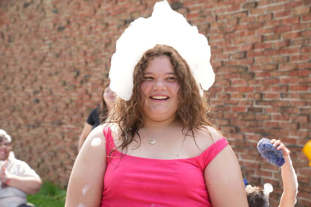 A smiling Summer School student in a bright pink top stands outside with wet hair and a large pile of bubbles resting on top of her head during Water Play Day.