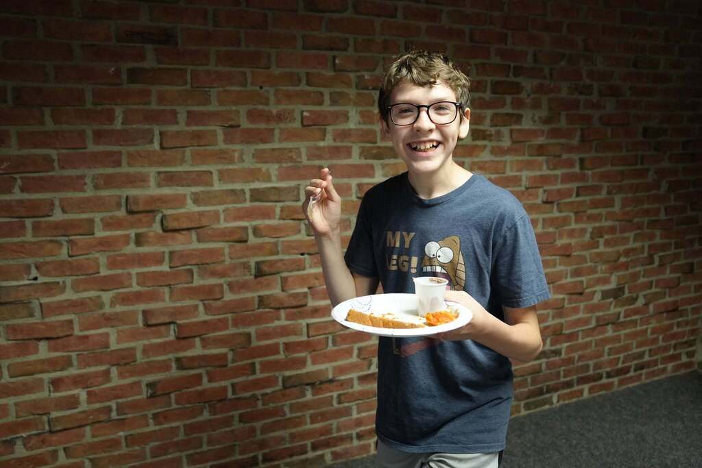 A smiling Summer School student wearing glasses holds a plate of food and a fork while sampling dishes during the World Cup Flavor event.