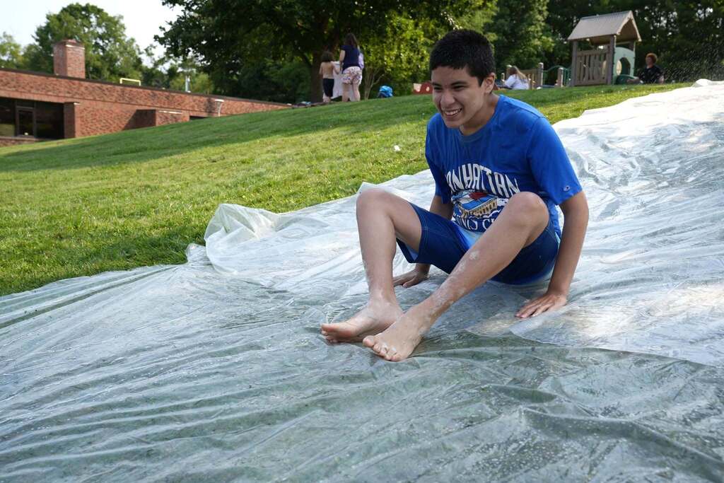 A smiling Summer School student sits at the bottom of a large outdoor slip and slide after sliding down the grassy hill during Water Play Day.