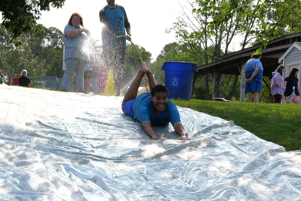 A Summer School student smiles while sliding headfirst down a large water-covered slip and slide as staff spray water from the top of the hill during Water Play Day.