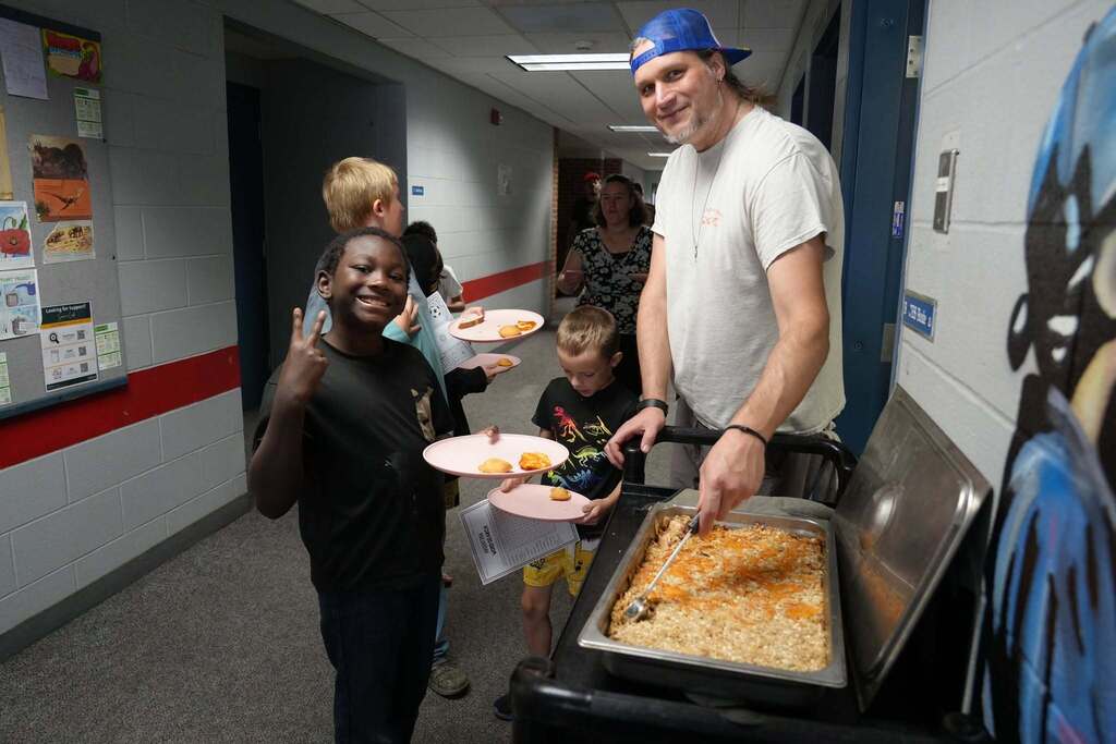 A smiling student holds a plate while another student receives a serving of food from a staff member during the World Cup Flavor event at Smith School.