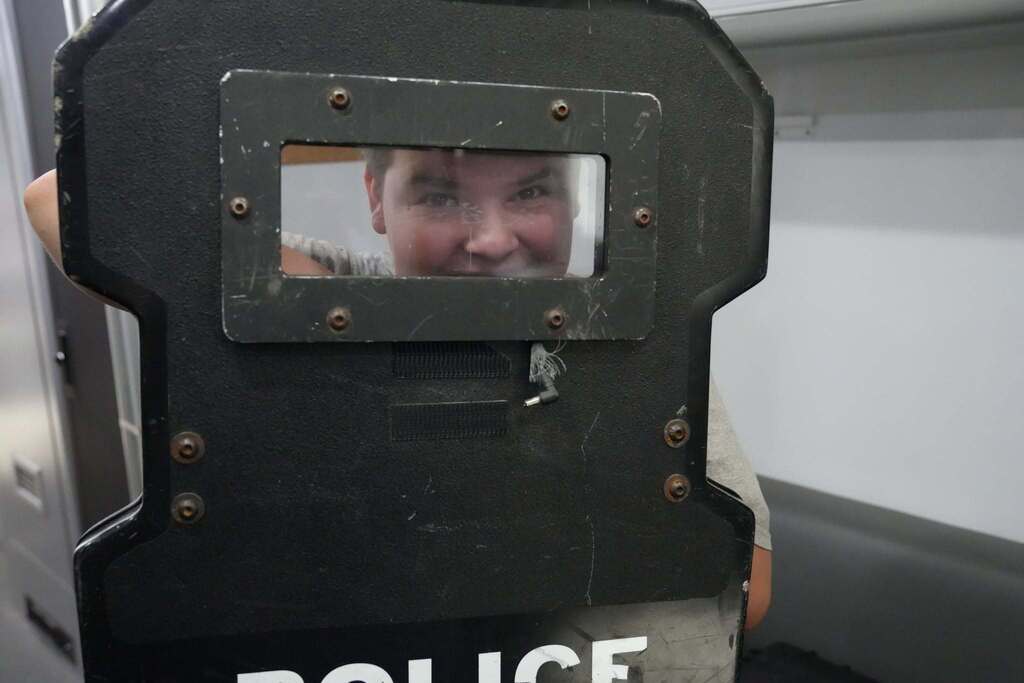 A student smiles through the viewing window of a police protective shield while exploring specialized law enforcement equipment.