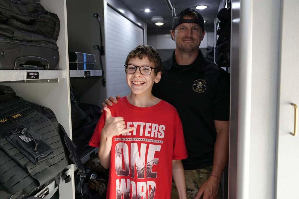 A smiling student gives a thumbs up hand gesture while standing with a police officer inside a specialized law enforcement vehicle.