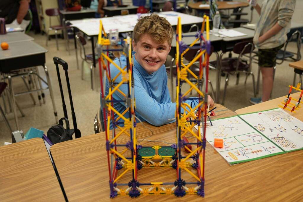 A smiling young man with blonde hair sits at a classroom table behind a tall, complex structure he built using red, yellow, and blue plastic construction rods, K'nex.