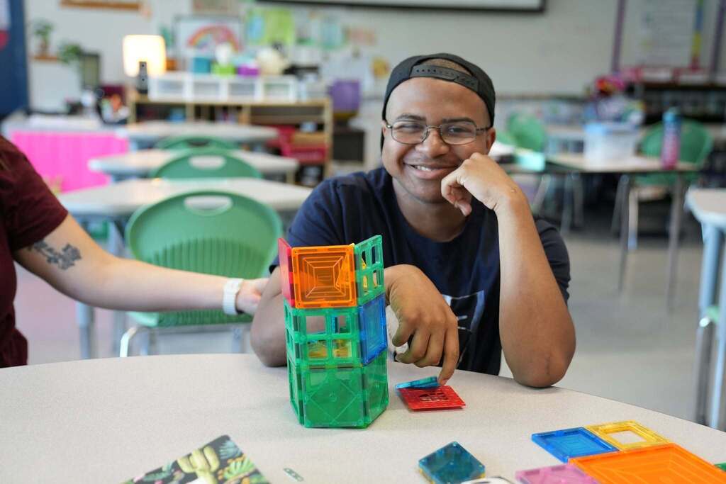 A smiling young person with glasses and a black baseball cap sits at a classroom desk, gesturing toward a colorful, hexagonal tower they built with transparent geometric magnetic tiles.