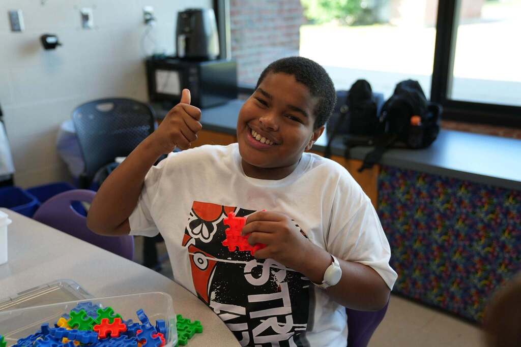 At a classroom table, a smiling young person in a graphic white t-shirt gives a thumbs-up with one hand while holding and working on interlocking red puzzle pieces.