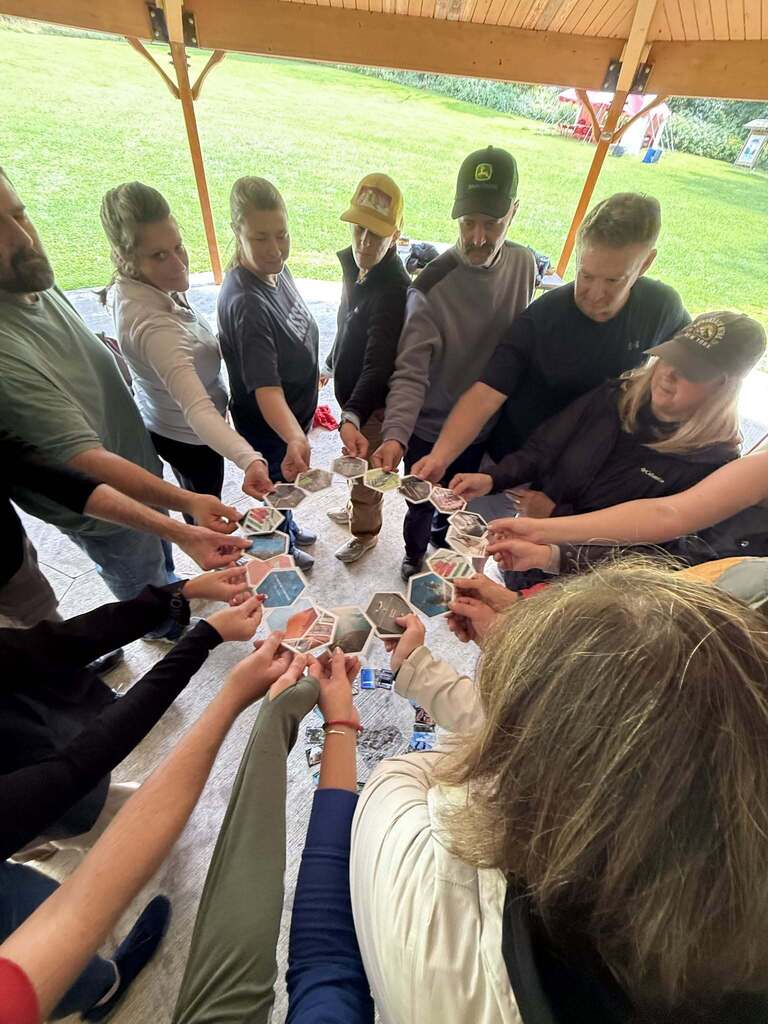 Eleven people stand in a circle under a wooden gazebo roof, holding small hexagonal photo cards and forming a large ring with their linked hands, as a group outdoor team-building activity.