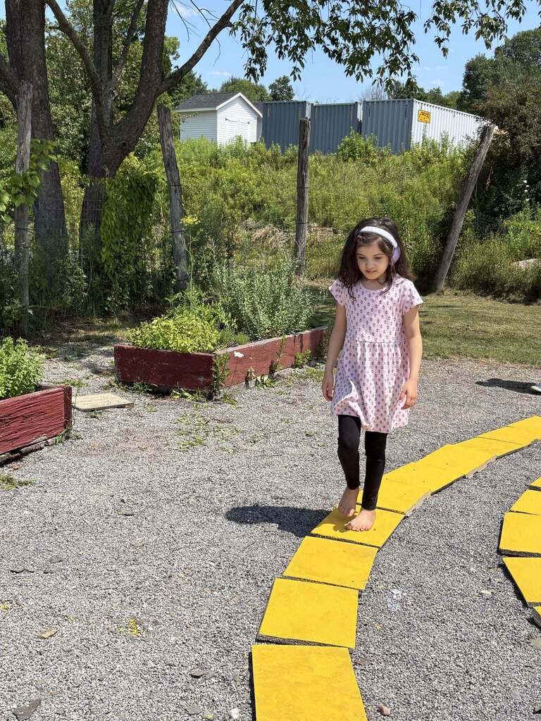 A young girl wearing a pink patterned dress and white headphones walks barefoot with focused balance along a curved path of large yellow stepping stones in a garden setting.