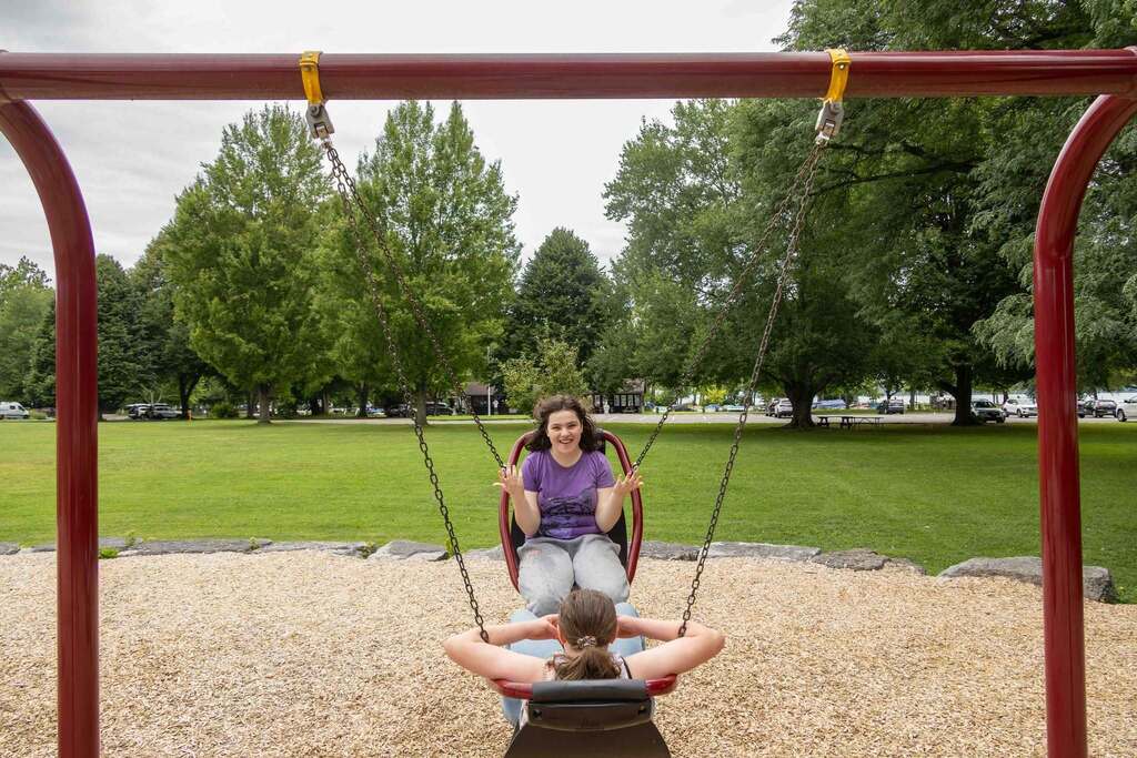 In a green park with large trees, two girls share an accessible swing seat; the girl facing forward in a purple shirt smiles with her hands raised in excitement.