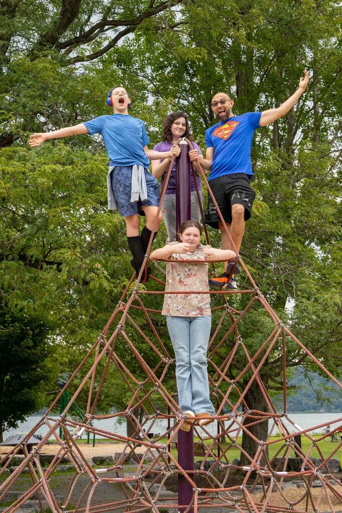 : A group of four people, three children and one adult man wearing a blue Superman t-shirt, climb and balance on a tall red rope pyramid structure in a grassy park, all with joyful expressions.
