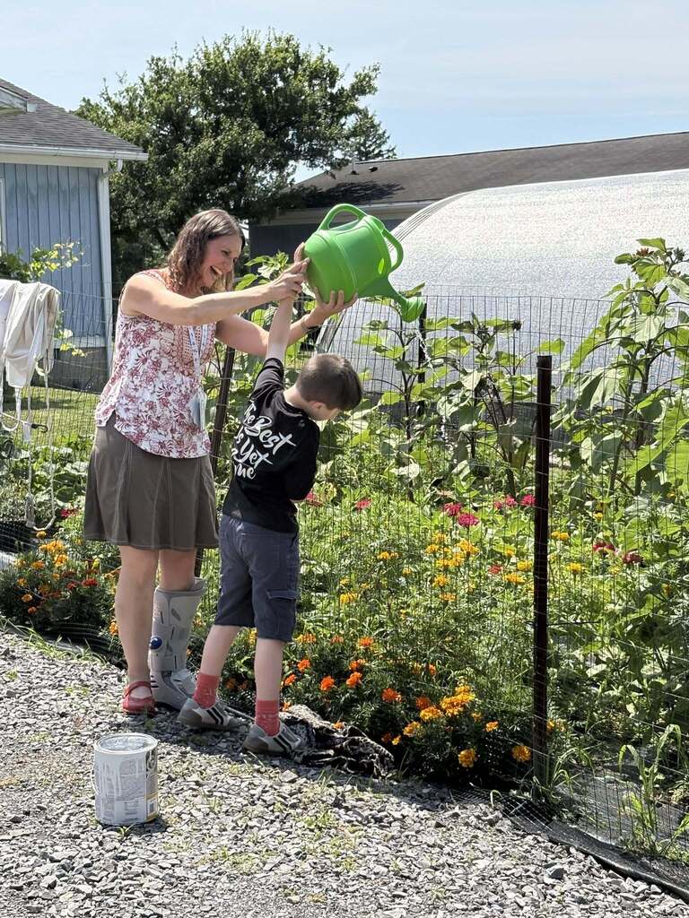 : A woman with a leg cast and a floral shirt lifts a large green watering can, assisting a young boy to pour water on blooming marigolds and green plants in a sunlit garden bed next to a greenhouse.