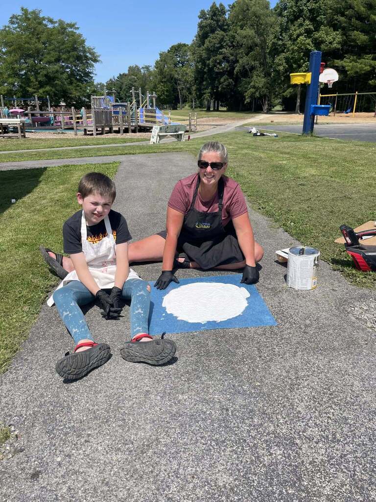 Two people are on a gravel path: a woman wearing sunglasses and a black apron sits next to a young boy in a white apron, and together they are painting a large white circle on a blue mat on the ground.
