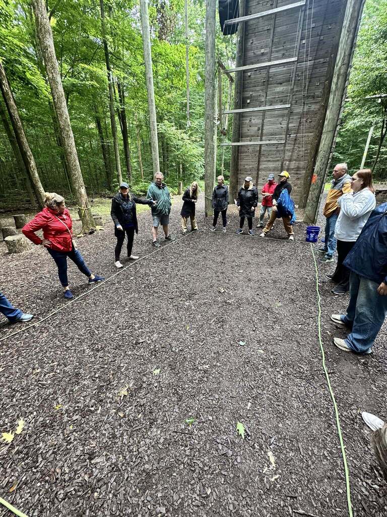 A group of about ten adults in casual clothing are standing in a large rectangle marked with green rope on a wood chip course in the forest, with a man addressing them near a tall wooden climbing wall, at the Hoffman Challenge Course.