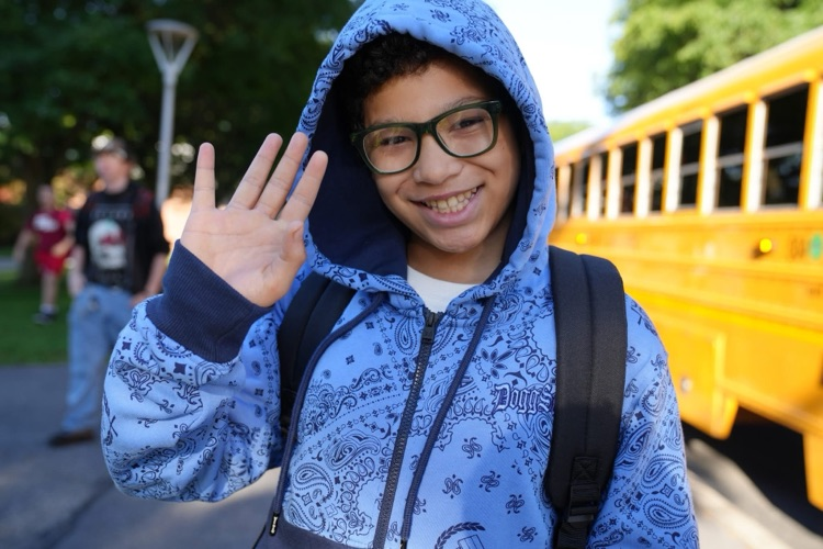 A boy wearing green glasses and a blue patterned hooded sweatshirt waves his hand at the camera while standing outdoors next to a yellow school bus.
