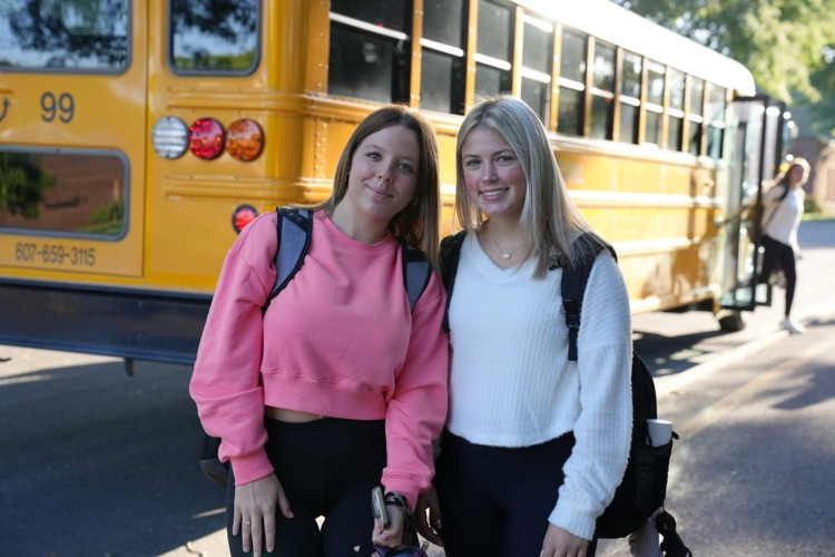 Two teenage girls smiling together in front of a yellow school bus, both wearing backpacks. One wears a bright pink cropped sweatshirt and the other wears a white sweater.
