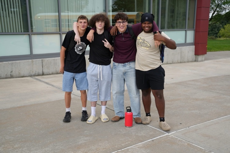 Four high school students standing outside P-TECH Academy with their arms around each other's shoulders, smiling for the camera. A red water bottle sits on the concrete ground in front of them.