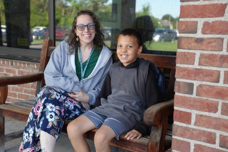 A woman with glasses, a grey zip-up hoodie, and floral pants sits next to a young boy in a dark hoodie and grey shorts on a wooden bench outside a brick school building. Both are smiling at the camera.