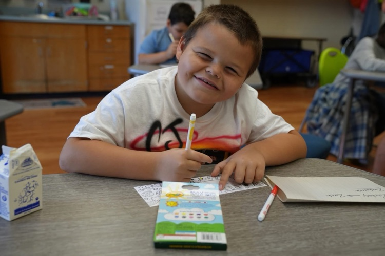 A young boy sitting at a classroom desk smiling while drawing with a marker. A box of Crayola markers, a small milk carton, and a notebook sit on the table in front of him.