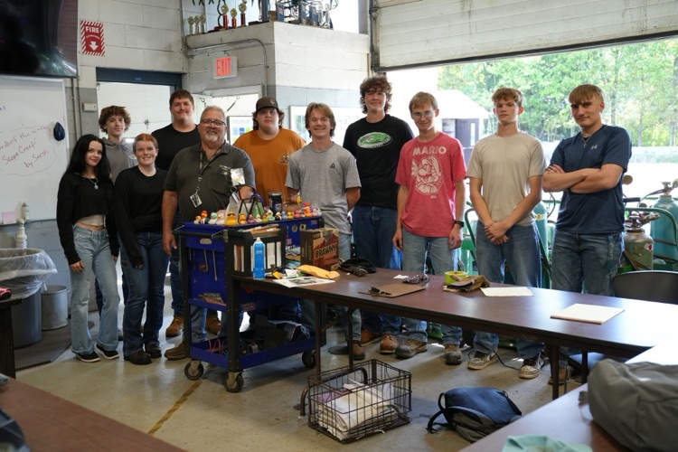 A group of welding students and their instructor posing together inside the welding classroom, standing around a large work table and a rolling tool cart.