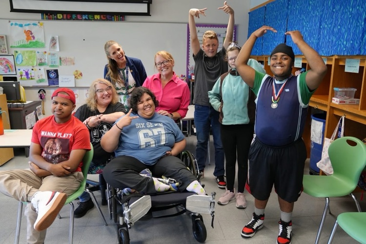 A group of students and teachers smiling together in a classroom. One student in a wheelchair strikes a peace sign pose, while another student in the front wears a medal and makes a crown shape over his head with his hands.