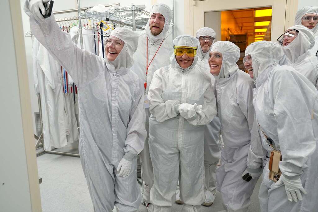 A group of visitors, with Commissioner Rosa in the middle, don cleanroom suits smile together while taking a selfie inside the Cornell NanoScale Science and Technology Facility.