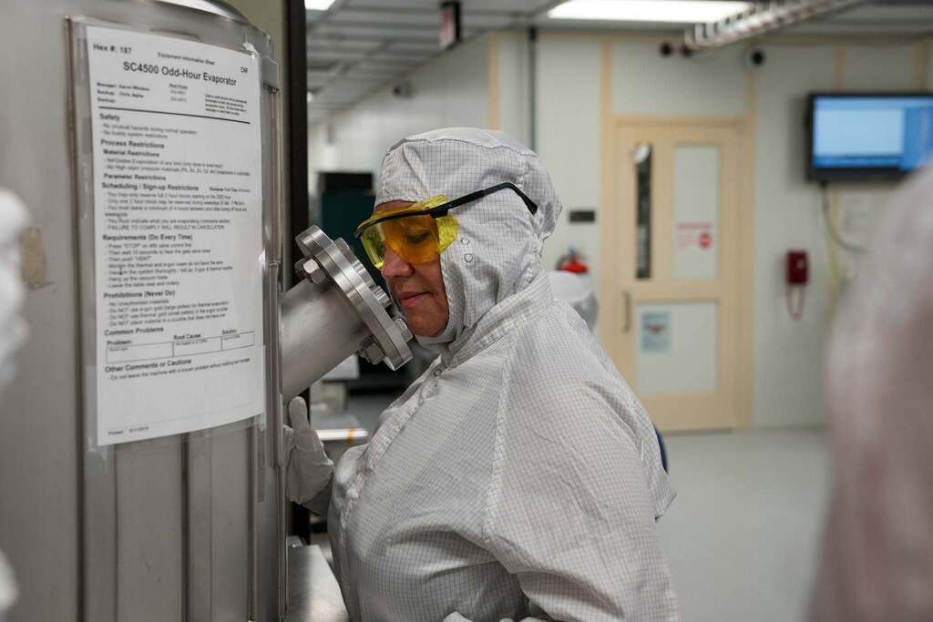 Commissioner Rosa wearing a cleanroom suit and yellow safety glasses looks closely into a piece of specialized equipment inside Cornell’s NanoScale Science and Technology Facility.