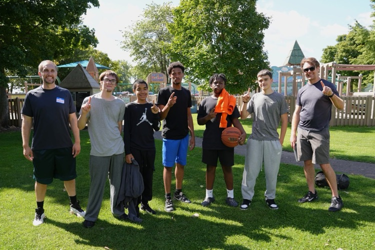 A group of RAS students and staff on a basketball court, with one holding a basketball and another an orange shirt. Their pose with ones raised shows great spirit and team camaraderie.