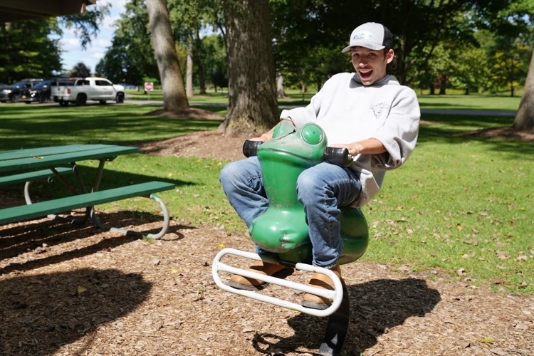 A joyful RAS student wearing a "Ford" cap leans back and laughs while riding on a frog on a spring on the playground, capturing a moment of picnic fun.