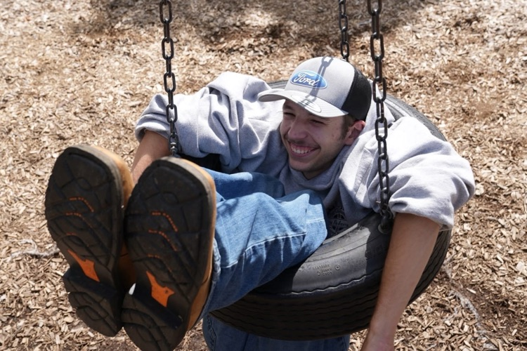 A joyful RAS student wearing a "Ford" cap leans back and laughs while inside a tire swing on the playground, capturing a moment of relaxed picnic fun.