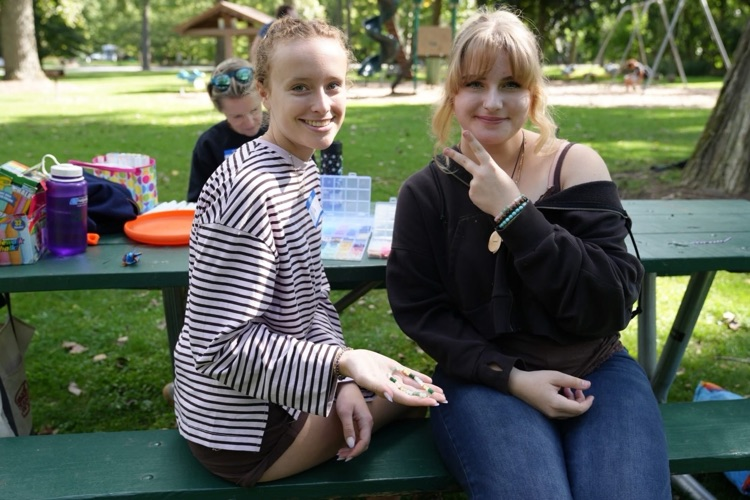 A close-up view of two RAS students at a picnic table, smiling. One holds a newly-made colorful beaded bracelet, showcasing one of the day's favorite creative activities.
