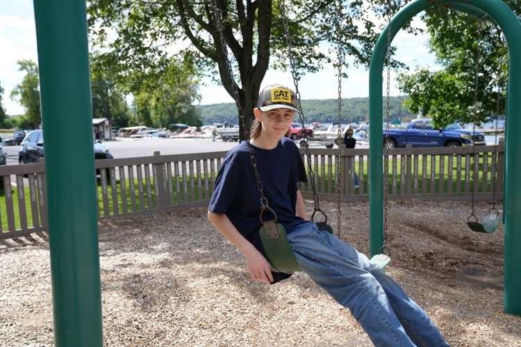 An RAS student in a "CAT" cap smiling happily on a traditional swing, representing classic playground fun near the water at Myers Park.