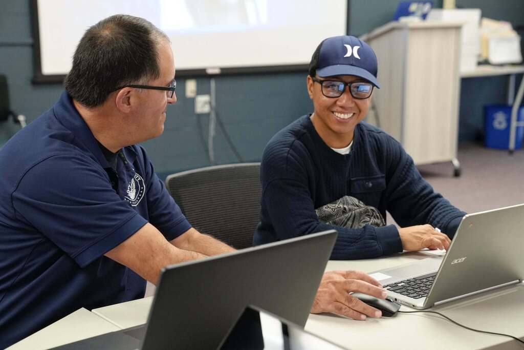 Tompkins County staff member sits beside a hiring event applicant, assisting him on a laptop as he smiles at the camera.