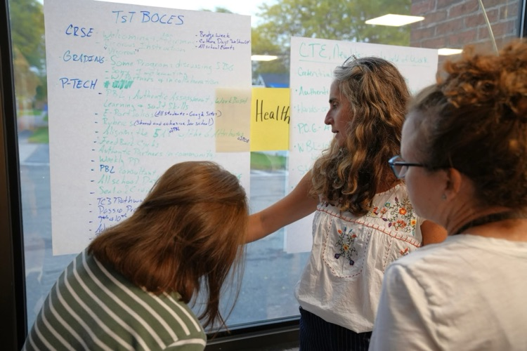 Three participants standing together by a window review handwritten notes about TST BOCES programs posted on the glass.