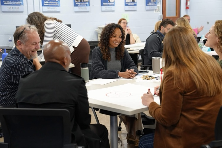A group of educators gathered around a table smiling and talking while writing notes on a large piece of chart paper.
