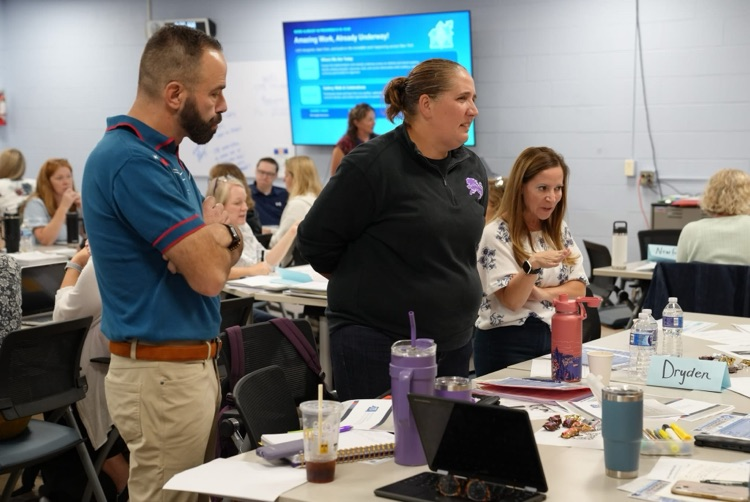 Three school representatives standing near a conference table labeled "Dryden," collaborating and discussing ideas.
