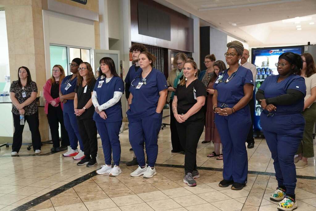 A crowd of people, including several Practical Nursing students in blue scrubs, stand observing the speakers and event during the ribbon-cutting ceremony.