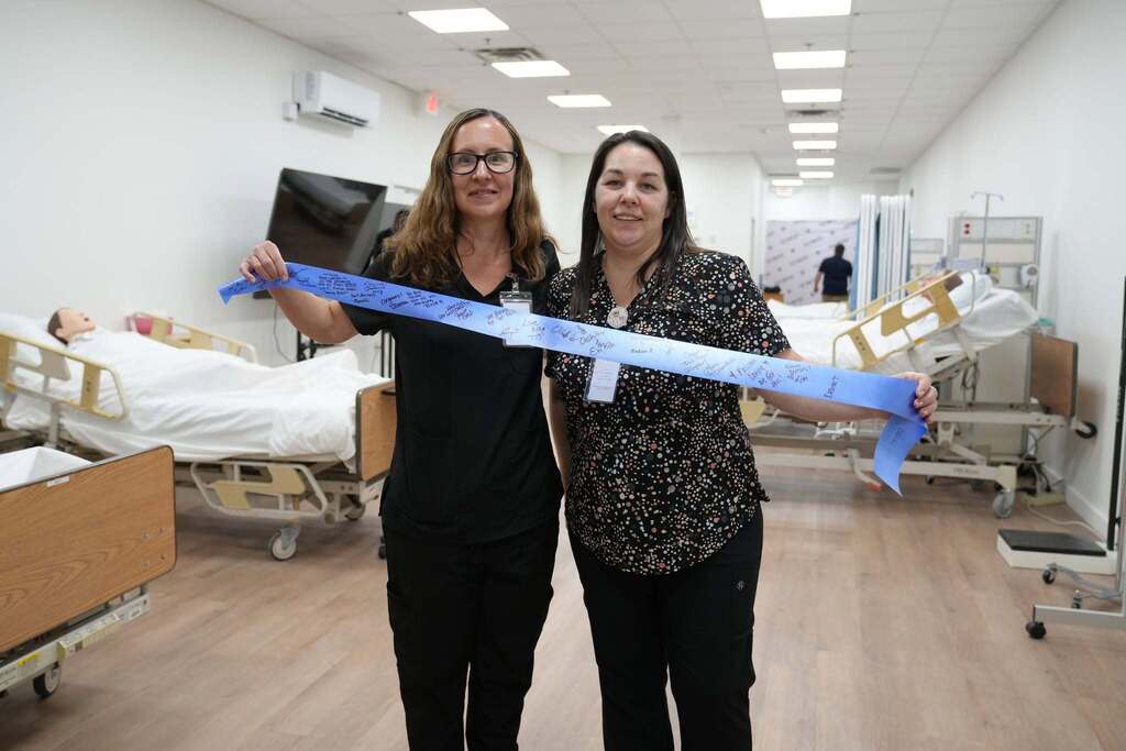 Two women in scrubs smile as they hold up the blue ceremonial ribbon covered in hand-written signatures. They are standing in the simulation lab surrounded by hospital beds