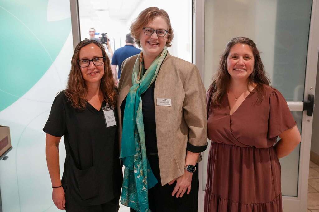 A group of three women smiling together near a glass entryway. The women are representing TST BOCES TC3. and One woman wears black scrubs, the center woman wears a tan blazer and a blue and teal scarf, and the third woman wears a maroon dress.