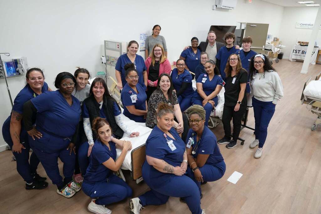 An overhead view of a group of Practical Nursing students in blue scrubs posing together in a simulation lab. They are gathered around a hospital bed with other medical equipment visible in the background.