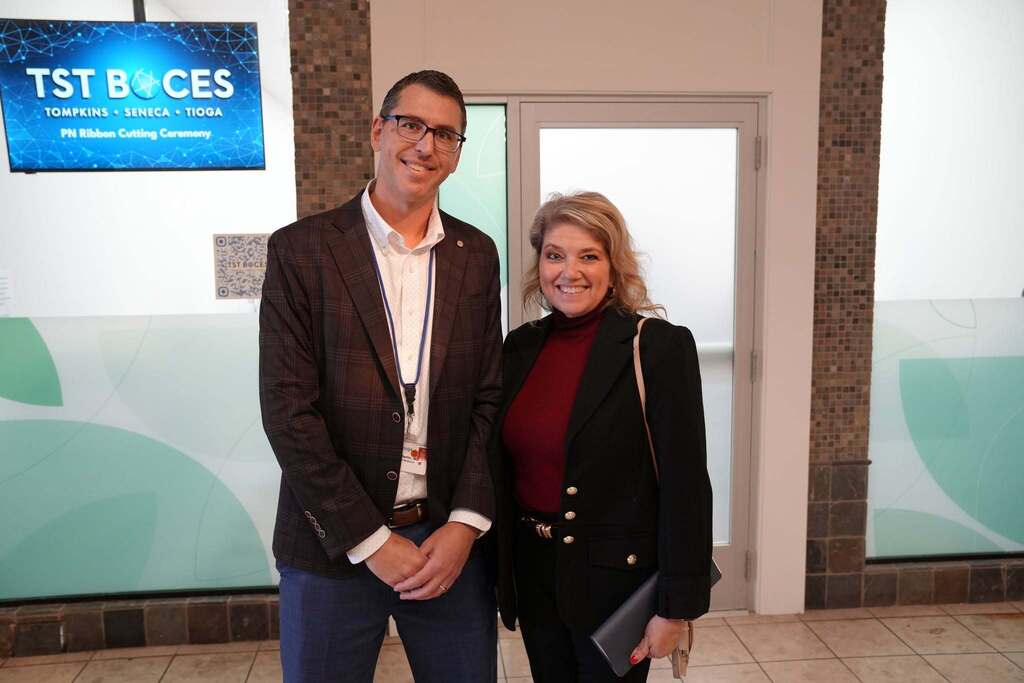A close-up photograph of Dr. Michael Berlin and Cindy Walter smiling together in the mall foyer following the ceremony. The TST BOCES screen is visible behind them