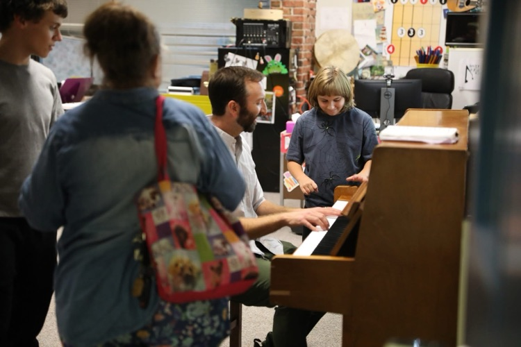 music teacher plays the piano with students in the background and one joins in on the piano with him to play