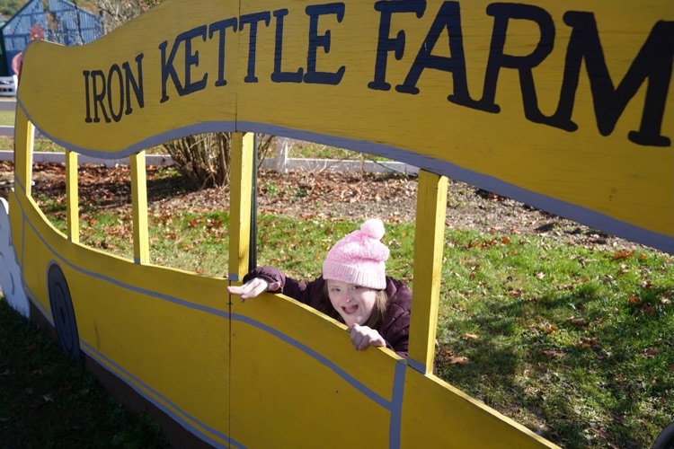 Student pokes her head through a wooden cut out in the shape of a yellow bus that says "iron kettle farm" on the top and smiles at the camera on class field trip