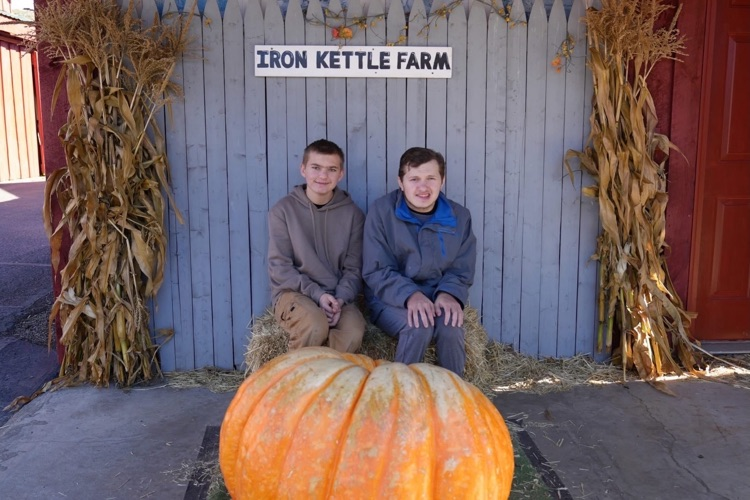 two students sit on hay bale behind a large pumpkin and and smile at the camera on class field trip