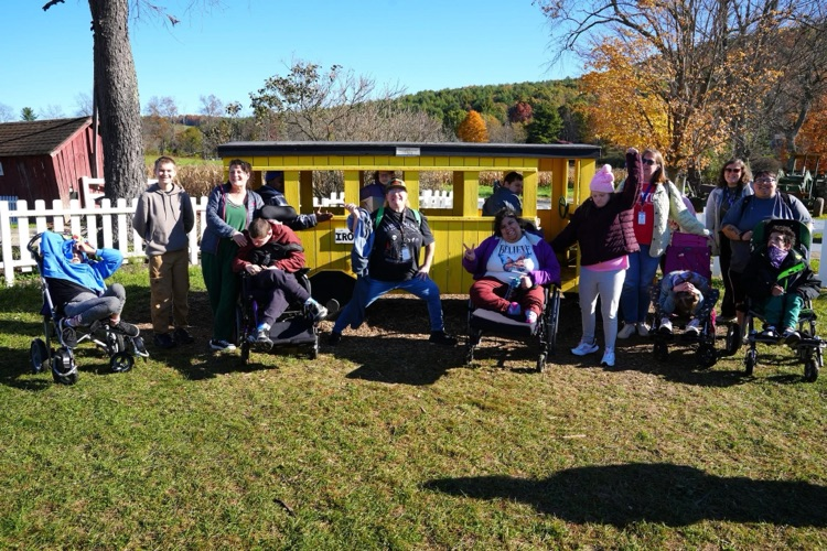 both class groups stand in front of a yellow wooden train and pose for photo and smile at the camera on class field trip