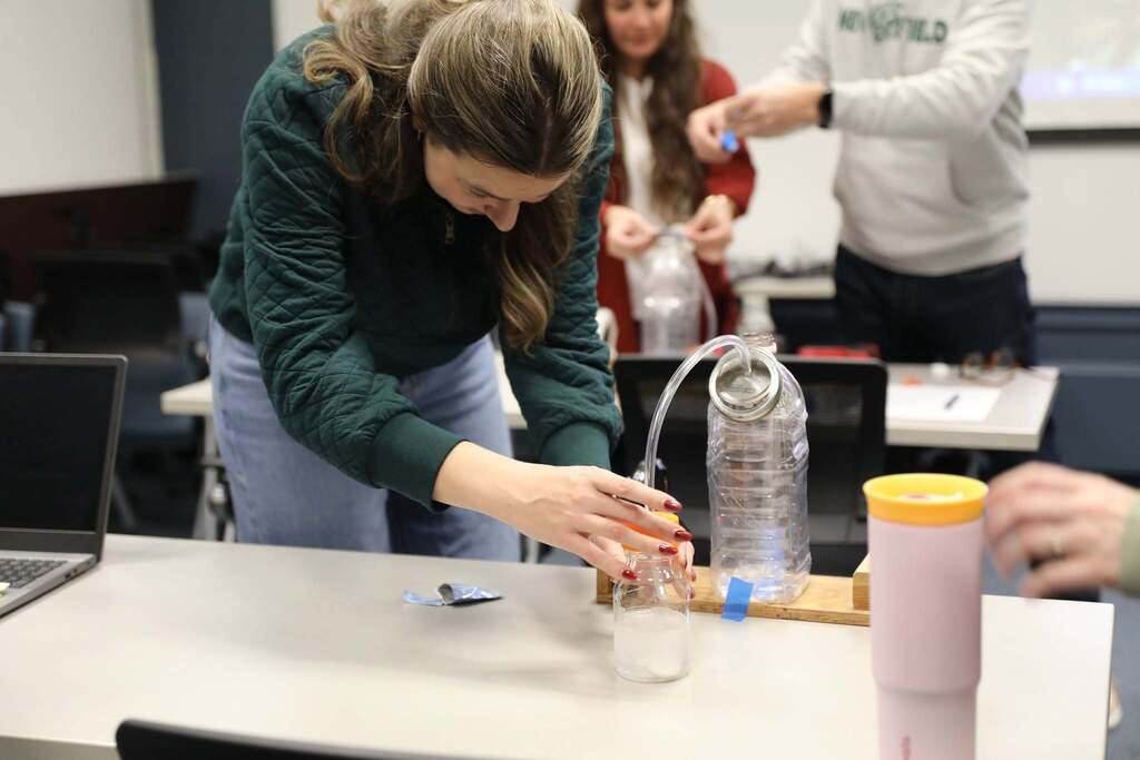 An educator leans over a table to carefully adjust plastic bottles, tubing, and liquid during a hands-on STEM experiment activity.