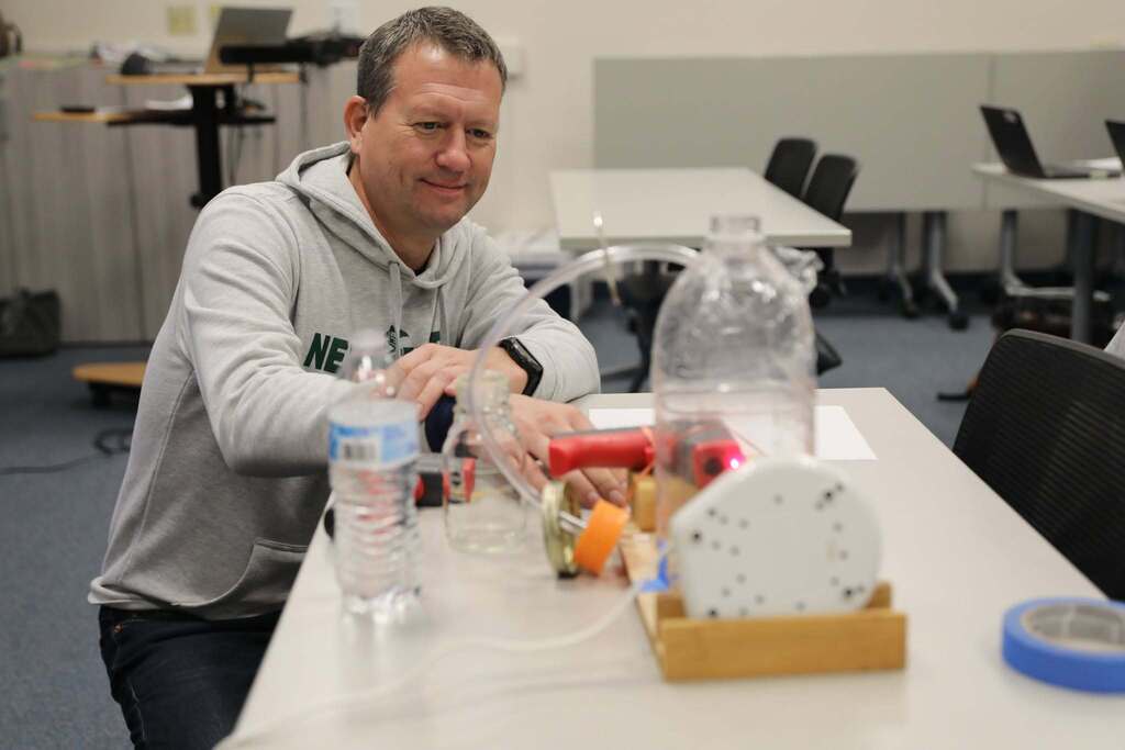 A man in a grey hoodie smiles while inspecting a hands-on science or STEM demonstration setup involving plastic bottles and tubing on a table.
