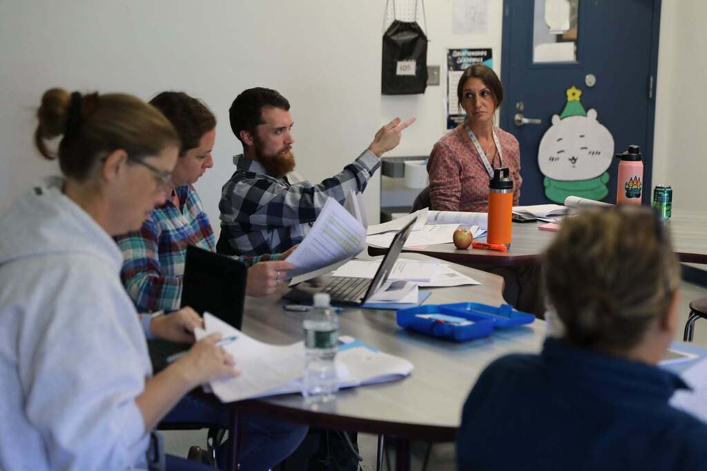 A man pointing during a group discussion at a round table surrounded by papers, notebooks, and laptops.