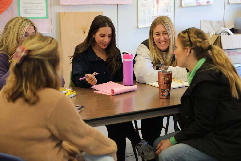 Four educators sit together around a table engaged in a small-group discussion, taking notes on a pink notepad during a professional learning activity.