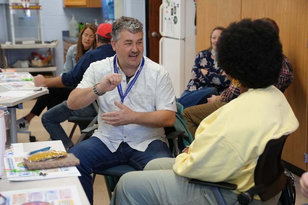 A male educator wearing a lanyard gestures enthusiastically while conversing with a colleague during a group session.