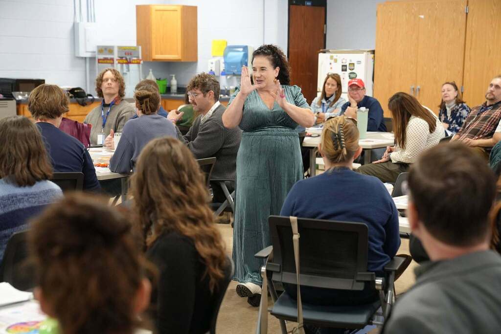 A facilitator wearing a green dress stands in the center of a classroom holding her hands up as she speaks, with seated educators facing her in a semi-circle during a workshop.